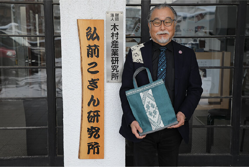 津軽こぎん刺しのスーツとネクタイ、ブローチ、バッグを身につける成田さん